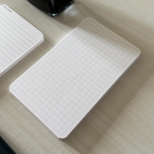 A side view of a set of index card-sized, note cards on a white table. The card on the left has a line with mini vertical dots layout with the text "Pockets + Pages by Turnip" at the bottom. The stack of cards on the right has a graph layout. A black ink bottle and small blue pen is placed nearby. Additional text on the bottom left in the image provides more details on the product including the size, material, and layout description.