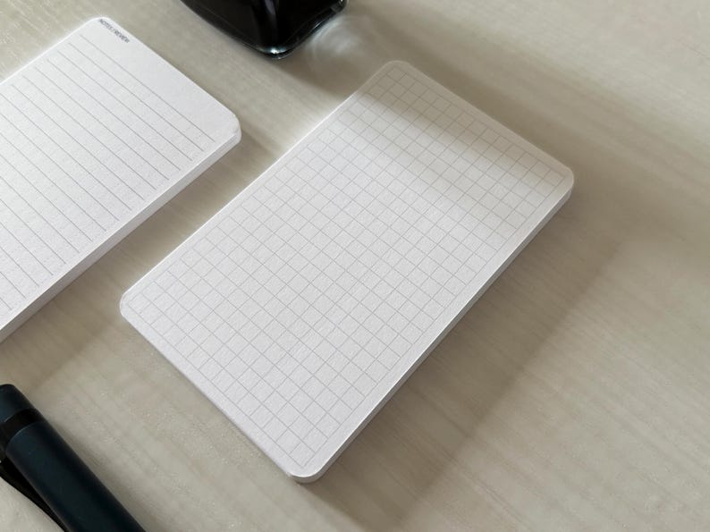 A side view of a set of index card-sized, note cards on a white table. The card on the left has a line with mini vertical dots layout with the text "Pockets + Pages by Turnip" at the bottom. The stack of cards on the right has a graph layout. A black ink bottle and small blue pen is placed nearby. Additional text on the bottom left in the image provides more details on the product including the size, material, and layout description.