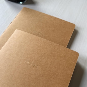 May include: Two brown notebooks with the words "Thoughts" and "notes" embossed on the covers. The notebooks are stacked on top of each other, with the top notebook slightly angled. The background is a light, neutral color.