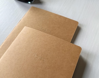 Può includere: Due quaderni marroni con le parole "Thoughts" e "notes" impresse sulle copertine. I quaderni sono sovrapposti, con quello superiore leggermente angolato. Lo sfondo è di un colore chiaro e neutro.