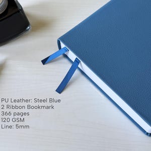 A blue PU-leather notebook sits on a white wooden desk, showing 2 different color ribbon bookmarks that are of similar shade to the cover: dark blue and navy blue. There is a fountain pen and ink bottle placed next to the notebook. Additional text in the image states that the notebook is a Steel Blue color, has 366 pages, 120gsm paper, and is 5mm line.