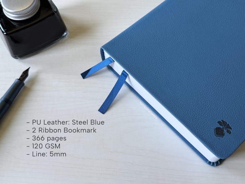 A blue PU-leather notebook sits on a white wooden desk, showing 2 different color ribbon bookmarks that are of similar shade to the cover: dark blue and navy blue. There is a fountain pen and ink bottle placed next to the notebook. Additional text in the image states that the notebook is a Steel Blue color, has 366 pages, 120gsm paper, and is 5mm line.