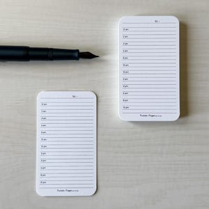 May include: Two white notepads with rounded corners, each with a lined schedule from 12 am to 10 pm. A black fountain pen lies across the top of the notepads. The text "to -" is at the top of each notepad.