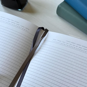An open notebook showing a clean, structured layout using solid lines with vertical dots, featuring an undated days, months, and weeks calendar at the top. Two PU-leather notebooks with green and blue covers are stacked nearby on a white desk. There is a black fountain pen on top of the notebook and an ink bottle nearby.