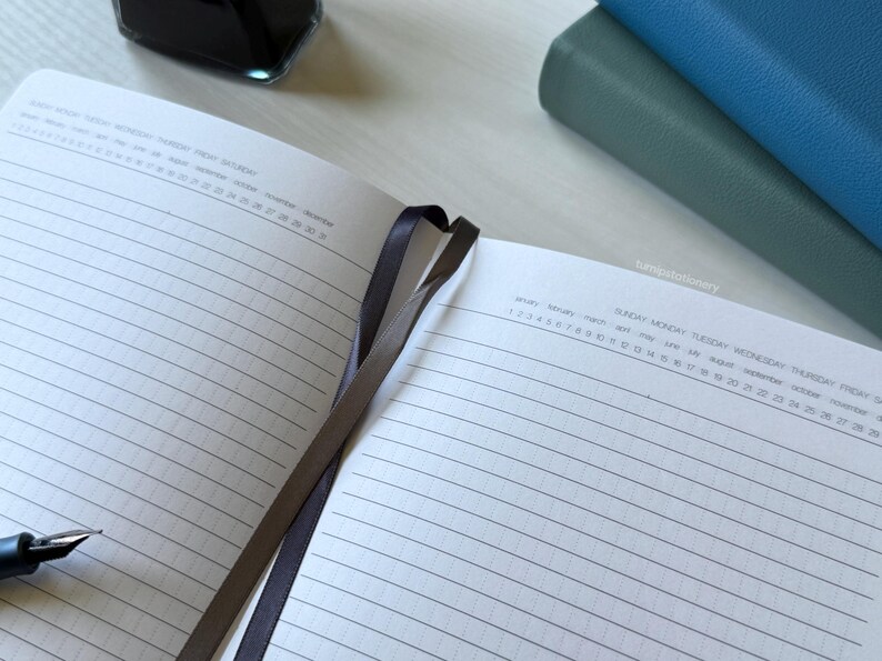 An open notebook showing a clean, structured layout using solid lines with vertical dots, featuring an undated days, months, and weeks calendar at the top. Two PU-leather notebooks with green and blue covers are stacked nearby on a white desk. There is a black fountain pen on top of the notebook and an ink bottle nearby.