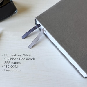 A gray PU-leather notebook sits on a white wooden desk, showing 2 different color ribbon bookmarks that are of similar shade to the cover: charcoal and dark gray. There is a fountain pen and ink bottle placed next to the notebook. Additional text in the image states that the notebook is a Silver color, has 366 pages, 120gsm paper, and is 5mm line.