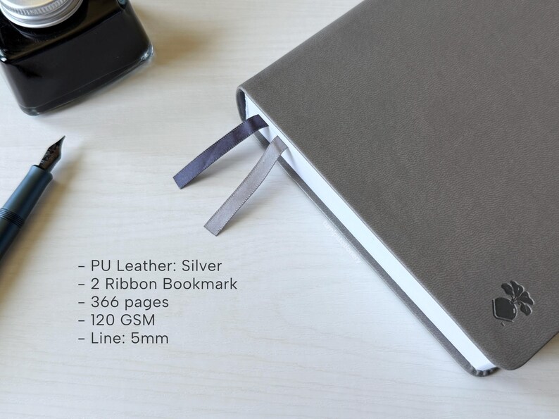 A gray PU-leather notebook sits on a white wooden desk, showing 2 different color ribbon bookmarks that are of similar shade to the cover: charcoal and dark gray. There is a fountain pen and ink bottle placed next to the notebook. Additional text in the image states that the notebook is a Silver color, has 366 pages, 120gsm paper, and is 5mm line.