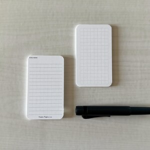 A set of business card-sized, note cards on a white table. The card on the left has a line with mini vertical dots layout with the text "Pockets + Pages by Turnip" at the bottom. The stack of cards on the right has a light graph layout. A small blue pen is placed nearby.