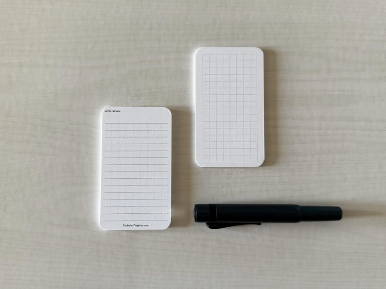 A set of business card-sized, note cards on a white table. The card on the left has a line with mini vertical dots layout with the text "Pockets + Pages by Turnip" at the bottom. The stack of cards on the right has a light graph layout. A small blue pen is placed nearby.