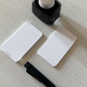 A side view of a set of business card-sized, note cards on a white table. The card on the left has a line with mini vertical dots layout with the text "Pockets + Pages by Turnip" at the bottom. The stack of cards on the right has a graph layout. A black ink bottle and small blue pen is placed nearby.