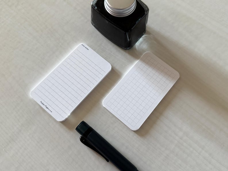 A side view of a set of business card-sized, note cards on a white table. The card on the left has a line with mini vertical dots layout with the text "Pockets + Pages by Turnip" at the bottom. The stack of cards on the right has a graph layout. A black ink bottle and small blue pen is placed nearby.