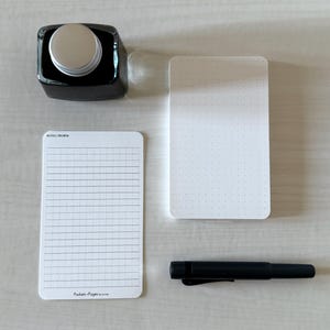 May include: An overhead shot of stationery items, including a black ink bottle with a silver lid, two white notepads, and a black pen. One notepad has a grid pattern, the other has a dot grid. The pen is lying horizontally.