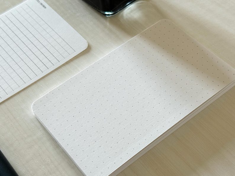 A focus view of a set of index card-sized note cards on a white table. The card on the left has a line with mini vertical dots layout with the text "Notes/Review" at the top left of the card. The stack of cards on the right has a dot grid layout. The dot grid card is focused in to show the mini dots. A black ink bottle and small pen is placed nearby.