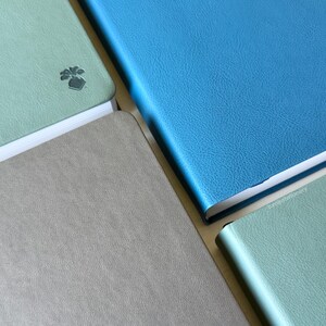 A collection of softcover PU-leather notebooks with covers in blue, green, and gray, arranged in a grid-like pattern. One cover shows a small embossed turnip design on the bottom corner.