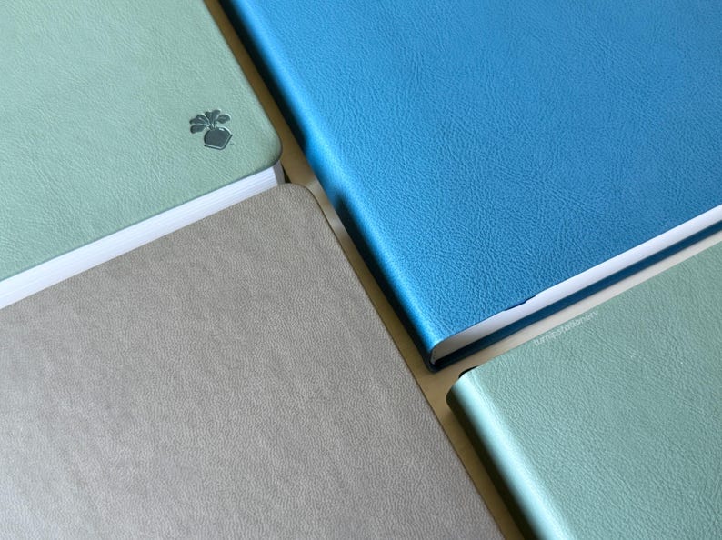 A collection of softcover PU-leather notebooks with covers in blue, green, and gray, arranged in a grid-like pattern. One cover shows a small embossed turnip design on the bottom corner.