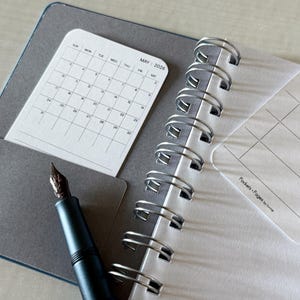 May include: Open notebook with a gray cover, spiral binding, and a pocket holding a May 2026 calendar. A black fountain pen with a brown nib rests on the open pages. The notebook has a white interior with lined pages.