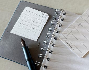 May include: Open notebook with a gray cover, spiral binding, and a pocket holding a May 2026 calendar. A black fountain pen with a brown nib rests on the open pages. The notebook has a white interior with lined pages.