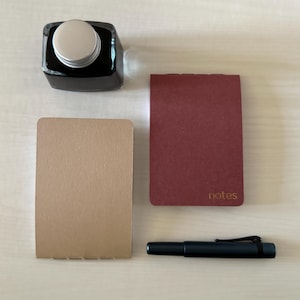 May include: An overhead shot of stationery items including a black ink bottle, two small notebooks, and a black pen. One notebook is tan, the other is burgundy with the word "notes" printed on it.