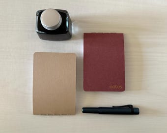 May include: An overhead shot of stationery items including a black ink bottle, two small notebooks, and a black pen. One notebook is tan, the other is burgundy with the word "notes" printed on it.
