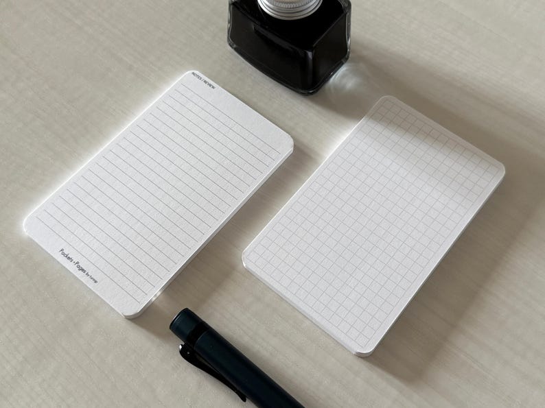 A side view of a set of index card-sized, note cards on a white table. The card on the left has a line with mini vertical dots layout with the text "Pockets + Pages by Turnip" at the bottom. The stack of cards on the right has a graph layout. A black ink bottle and small blue pen is placed nearby.