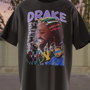 May include: Black t-shirt with a graphic print of Drake. The graphic features a collage of images of Drake, including a close-up of his face, a photo of him wearing a blue jacket, and a photo of him with a colorful background. The text "DRAKE" is printed in purple letters on the shirt.
