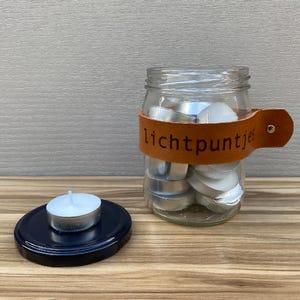 Op de afbeelding: Een glazen pot met een bruin leren bandje met de tekst "Lichtpuntjes" gevuld met witte theelichtjes. Een enkel theelichtje ligt op een zwart deksel.