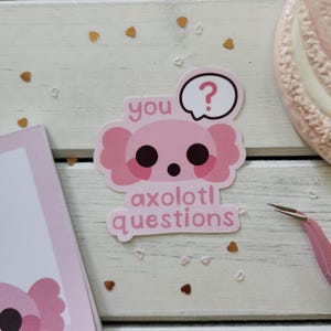 May include: A pink axolotl sticker with the text "you axolotl questions" and a speech bubble with a question mark. The sticker is on a white wooden surface with heart-shaped confetti.