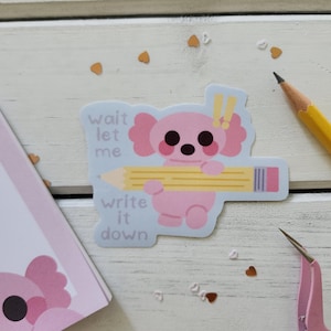 May include: A pink koala sticker holding a yellow pencil with the text "wait let me write it down" on a light blue background. The sticker is surrounded by small heart-shaped confetti and a yellow pencil.