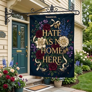 Könnte beinhalten: Eine rechteckige Gartenflagge, die an einer schwarzen Metallhalterung hängt, mit dunkelblauem Hintergrund, tiefroten Rosen, weißen Blumen, blauen Ritterspornen und zwei kleinen Vögeln. Der Text lautet HATE HAS NO HOME HERE in goldener Schrift.