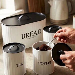 Könnte beinhalten: Ein Set aus weißen Metall-Küchendosen mit schwarzen Deckeln und Griffen. Der große ovale Brotkasten ist mit BREAD HOME STORAGE beschriftet. Zwei kleinere zylindrische Dosen tragen die Aufschriften TEA und COFFEE, wobei Kaffee enthalten ist.