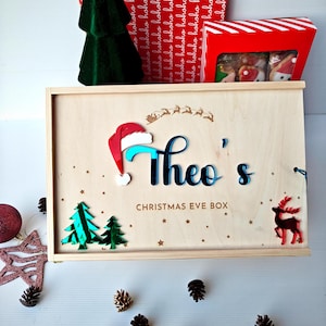 May include: A personalised wooden Christmas Eve box, featuring the name "Theo's" in blue, a Santa hat, and festive decorations. The box is surrounded by pine cones, with a green tree and gift boxes in the background.