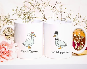 May include: Two white ceramic mugs with whimsical goose illustrations. One mug features a goose in a bridal veil, the other a goose in a top hat and bow tie. Text reads "Mrs. Silly goose" and "Mr. Silly goose".