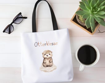 Könnte beinhalten: Weiße Tragetasche mit schwarzen Griffen. Sie zeigt eine Aquarellillustration eines Otters auf einem Felsen, darüber steht das Wort "Otterklasse". Eine Sonnenbrille, eine Sukkulentenpflanze und eine Kaffeetasse sind ebenfalls zu sehen.