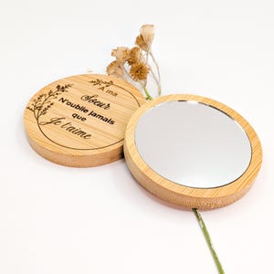 Puede incluir: Un espejo compacto redondo de bambú con una superficie reflectante plateada. El espejo está junto a una tapa de bambú con texto grabado y diseño floral. El texto dice "A ma soeur, N'oublie jamais que je t'aime."