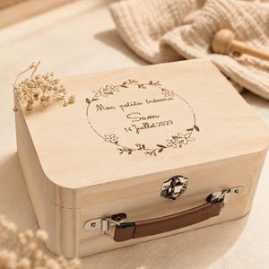 Peut inclure: Une boîte en bois clair avec une poignée et un fermoir en métal. La boîte présente un motif de couronne florale avec le texte "Mes petits trésors Sam 14 Juillet 2023" gravé sur le couvercle. La poignée est marron.