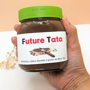 Peut inclure: Un pot de pâte à tartiner au chocolat et aux noisettes avec un couvercle vert. L'étiquette indique "Future Tata" en lettres rouges, avec une illustration d'une tranche de pain tartinée, des noisettes et un couteau. Le texte "Attention, câlins illimités à partir de Mars 2026" est également présent.
