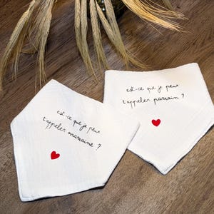 K&ouml;nnte beinhalten: Zwei wei&szlig;e Baby-L&auml;tzchen mit dem Text "est-ce que je peux t'appeler marraine?" und "est-ce que je peux t'appeler parrain?" in schwarzer Schrift, jedes mit einem kleinen roten Herz. Die L&auml;tzchen liegen auf einer Holzoberfl&auml;che.