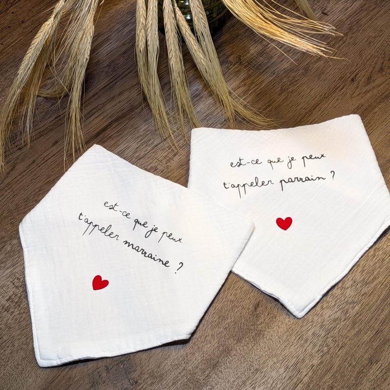 K&ouml;nnte beinhalten: Zwei wei&szlig;e Baby-L&auml;tzchen mit dem Text "est-ce que je peux t'appeler marraine?" und "est-ce que je peux t'appeler parrain?" in schwarzer Schrift, jedes mit einem kleinen roten Herz. Die L&auml;tzchen liegen auf einer Holzoberfl&auml;che.