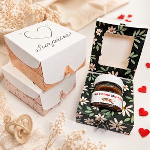 Peut inclure: Des boîtes cadeaux empilées, l'une ouverte révélant un pot avec un couvercle blanc et une étiquette "Je t'aime Maman". Les boîtes ont un cœur et le mot "Surprise!". La boîte ouverte a un motif floral.
