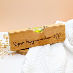 Puede incluir: Un nivel de madera con el texto "Super Papy en action" y un gráfico de un puño. El nivel tiene un vial transparente con líquido amarillo. El nivel está hecho de madera clara.