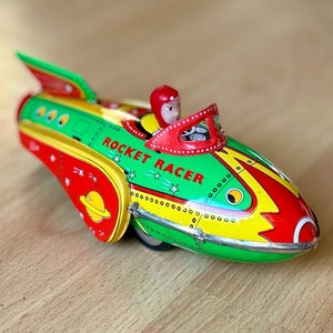 May include: A green, yellow, and red toy rocket ship with a wind-up key. The rocket has a small figure of a person in the cockpit and the words "Rocket Racer" printed on the side.