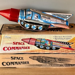 May include: A vintage tin toy rocket ship with a red nose and blue and white details. The toy is labeled "Space Commander" and features sparking lights and friction powered movement. The toy is displayed on its original box.