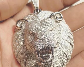 May include: A silver-colored lion pendant with a detailed design. The lion's head is covered in small, sparkling stones, with an open mouth revealing teeth. The pendant has a loop at the top for a chain.