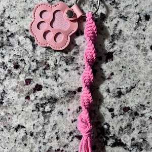 Op de afbeelding: Een roze sleutelhanger met een pootafdruk en een macramé kwastje. De pootafdruk is gemaakt van roze imitatieleer met een lichtere roze binnenkant en een zilveren sleutelring. Het kwastje is gemaakt van roze touw.