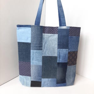 Puede incluir: Un bolso tote patchwork hecho de varios tonos de mezclilla. El bolso tiene forma rectangular con una abertura ancha y dos asas largas. Las piezas de mezclilla están cosidas en un patrón geométrico, creando un diseño único y texturizado. Adecuado para llevar comestibles o artículos personales.