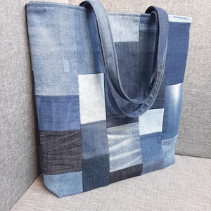 Puede incluir: Un bolso tote de mezclilla patchwork en varios tonos de azul. El bolso tiene forma rectangular con dos correas largas para llevarlo. Las piezas de mezclilla están cosidas en un patrón geométrico.