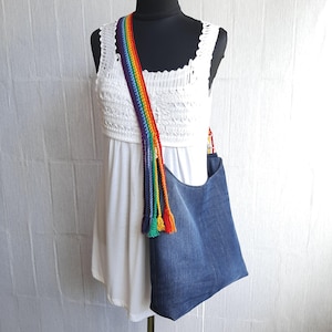May include: A denim tote bag with a rainbow-colored strap and tassels. The bag is a medium blue color and has a simple, casual design. The strap is made of a woven material in a rainbow pattern.