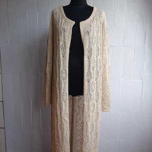 May include: A long, open-front cardigan in a light beige color. The cardigan features an intricate open-knit design with a repeating geometric pattern. The sleeves are long, and the cardigan extends to the ankles. The garment is displayed on a black mannequin.