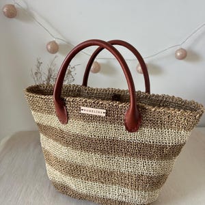 May include: Woven tote bag with brown and beige horizontal stripes. The bag has two brown leather handles and a small rectangular tag that reads "BOHELINE". The bag is designed for carrying items.