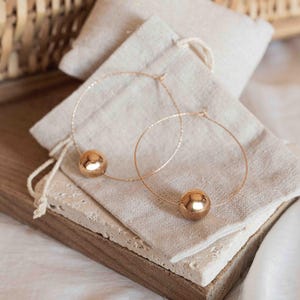 May include: A pair of rose gold hoop earrings, each featuring a single polished, spherical bead threaded onto a thin, textured wire hoop. The earrings are displayed resting on a small, light-colored fabric pouch, highlighting the metallic finish.